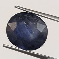 Oval faceted blue sapphire held with tweezers against a plain background, showing the front of the gemstone with visible facets and inclusions.