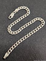 Silver chain bracelet laid out on a dark surface displaying curb link chain and lobster clasp.