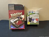 Front view of Deluxe Yahtzee Travel Edition box and GOLO! Golf Dice Game box side by side on beige background.