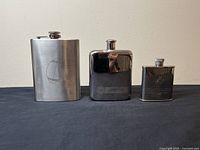Three vintage stainless steel hip flasks arranged side by side, showing different finishes and visible brand logos. The tallest is unbranded brushed steel with faint circular logo, the middle one is polished with Jameson branding, and the smallest is polished with Rémy Martin branding.