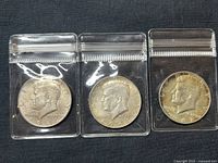 Three Kennedy Half Dollar coins in protective sleeves showing obverse side with profile of John F. Kennedy, dated 1964 and 1967.