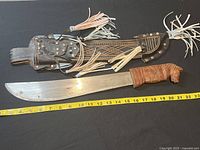 Full view of machete with measuring tape showing approximately 21 inches blade length, solid carved wood horse-shaped handle wrapped in leather, beside decorative leather sheath with brass studs and leather tassels.