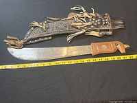 Full view of machete with decorative carved wooden horse handle, inset coin, and matching leather sheath with tassels and brass studs, measuring about 23 inches in length