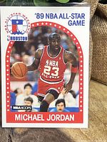 Front of 1989 NBA Hoops Michael Jordan basketball card showing him in All-Star uniform with number 23, holding basketball, in game action during 1989 NBA All-Star Game in Houston.