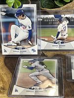 Five Shohei Ohtani baseball cards in protective sleeves arranged on a surface with plant background. Cards show Ohtani with LA Dodgers uniform in various baseball action poses.