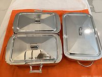 Two silver plated brass rectangular covered dish holders on orange cloth, one with a broken leg, shiny and reflective.