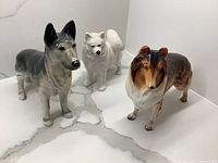 Front and angled view of the three dog figurines on a white marble surface, showing their details and colors.