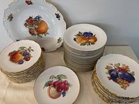 Large lot of Bavarian fine china dishes displayed, stacked in groups, showing fruit designs and gold trim on plates and platter.