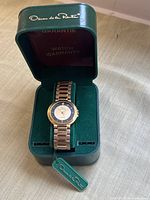 Oscar de la Renta branded green watch box containing the wristwatch on a green velvet cushion, showing warranty booklet inside lid and original tag attached to the watch bracelet.