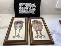 Three art pieces displayed together: signed Haida metal relief by Clarence Wells on black backing and two framed owl paintings in wooden frames on a white surface.