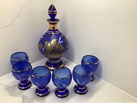 Full set photo showing one cobalt blue decanter with stopper and six matching glasses with gold etched floral designs and gold trim on a white marble surface.