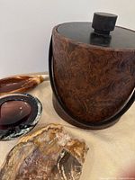 Photo showing leather tooled ice bucket with black lid and handle, plus three ashtrays around it on cream cloth.