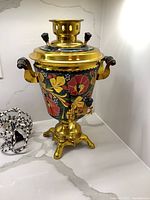 Full front view of vintage electric samovar showing gold tone metal body with hand painted red and yellow floral design, black handles, and spigot.