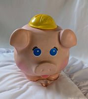 Front angled view of pink piggy bank wearing yellow hard hat showing facial features and shape
