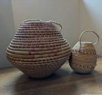 Two handwoven baskets placed side-by-side on a wooden surface. Left basket larger with round bulbous shape and handle on top. Right basket smaller, beaded as decoration and single loop handle on top.
