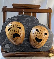 Full view of the two carved masks on dark stone plaque with rough edges and wooden easel stand behind.