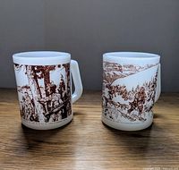 Two white Federal Glass Company mugs side by side on wooden surface, detailed brown illustrations of explorers and landscapes on each.
