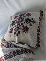 Single pillow case shown with geometric floral cross stitch patterns in browns and reds on beige fabric with fringed edges, draped to show texture and embroidery details.