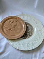 Two commemorative pottery plates: a terracotta-colored Fiesta Ware plate featuring a potter at the wheel relief stacked on a larger white Wedgwood plate with embossed floral border and central transferware image.