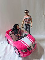Ken and Raquelle dolls posed with pink convertible car; Ken standing wearing striped shirt and sunglasses, Raquelle seated in car.