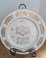 Front view of commemorative plate on stand showing gold decorative foliage, crossed flags, and text commemorating Apollo 11 moon landing July 20, 1969.