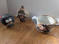 Three pottery items on a wooden surface: two piggy banks (one black, one beige) and one Eakin shaving cup with floral decoration.