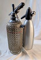 Photo of two soda syphon bottles: Sparklets soda syphon with woven metal mesh and iSi brushed aluminum soda syphon, standing side by side on a plain surface.