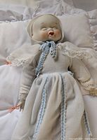 Front view of doll showing happy face, dressed in white with blue trim and lace sleeves, bonnet on head, laying on white ruffled bedding.