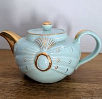 Front side of aqua blue vintage teapot showing scalloped design, gold accents on circular embossed element, and gold on spout and lid knob.