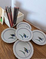 Five small round plates displayed in a semicircle showing kingfisher bird designs, accompanied by chopsticks leaning against boxes.