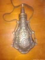 Front view of antique bronze powder flask with intricate embossed designs and leather strap attached, showing full height and nozzle.