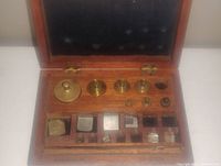 Antique wooden box containing a collection of brass and metal weigh scale weights, arranged in designated slots with a hinged lid open.