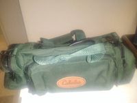 Green Cabela's fishing tackle bag shown from the side with handles and shoulder strap, oval brown logo patch visible.