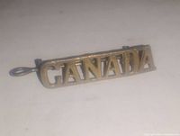 Close-up image of the vintage metal badge pin with the word 'CANADA' in raised gold-tone letters, showing the front and side details including the pin clasp.
