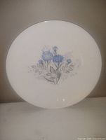 Photo of a white collectible ceramic plate with a blue floral pattern at the center, showing the entire front face of the plate.