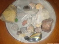 Assorted gems and minerals laid out in a round dish, showing clear quartz points, tiger's eye polished stone, and various other rough and polished stones.