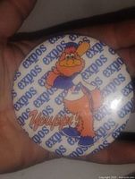 Close-up photo of a vintage Montreal Expos Youppi! button pin held in hand, showing the mascot with baseball bat and repeated 'expos' text in background.
