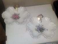 Two white porcelain decorative plates shaped like flowers with scalloped gold-trimmed edges, featuring pink rose and purple violet floral designs, displayed on a table.