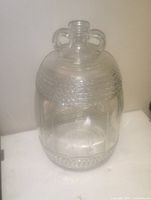 Clear glass vintage bottle with two handles and textured horizontal and vertical patterns, standing on a white surface under natural lighting.