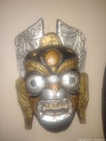 Front view of the handmade mask showing metal overlay on wooden base with gold and silver tones, intricate facial features including eyes, nose, mouth, ears with textured details.
