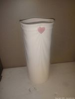 Tall white ceramic vase with smooth glazed finish and a small pink heart decoration on the front near the top. The top rim has a wavy design.