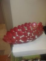 Photo showing a large bowl made of circular linked metal discs with a red finish, placed on a white surface against a light background.