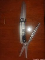 Photo of a silver multi tool open with knife blade, scissors, and another tool extended on wooden surface.