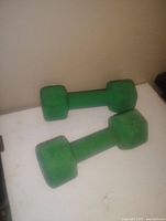 Pair of green neoprene dumbbells placed on white surface against beige wall, showing hexagonal ends and overall condition.