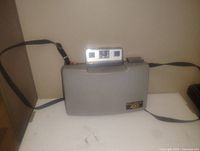 Vintage Polaroid PO420 420 instant camera in its grey carrying case with shoulder strap, placed on surface against a wall.