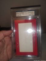 Photo of the PSA graded and authenticated encased autographed item with red background and certification label visible.