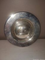 Photo shows a round silver plated wall plate with a reflective surface and a central embossed design.
