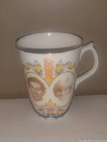 White ceramic cup featuring oval portraits of a man and woman with royal insignia and decorative detailing, silver rim around top.