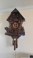 Front view of carved wooden clock with chalet roof, trees, figurines and pinecone weights hanging by chains
