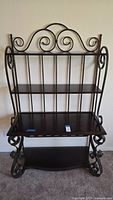 Front view of the bakers rack showing three wood shelves and decorative scroll metalier frame with curved accents on top and sides.